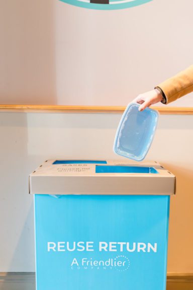 A hand dropping the friendlier plastic container into a reuse and return friendlier bin